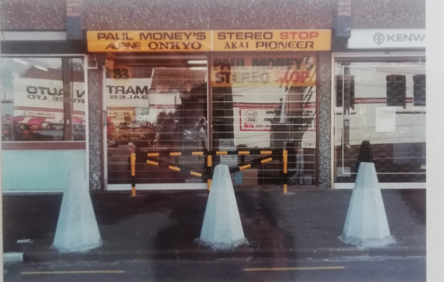 Storefront of a shop with large glass windows and signs above, blocked by a yellow-and-black striped barrier and three blue concrete traffic cones in the street.