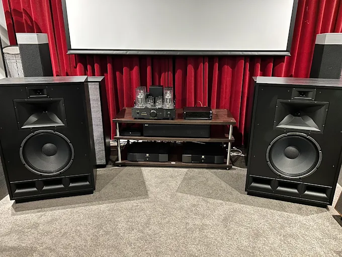Home theater setup with two large floor-standing speakers flanking a wooden equipment rack in front of red curtains and a projector screen